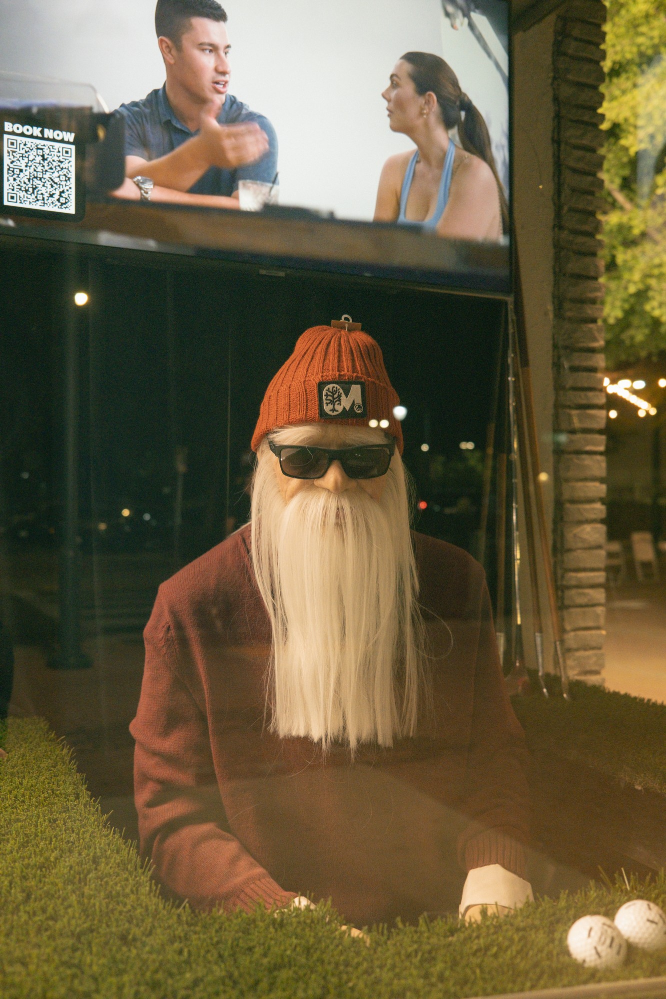 Real life Mulligan's mascot mannequin sitting in storefront window on synthetic turf with beanie, beard, sunglasses, and golf balls